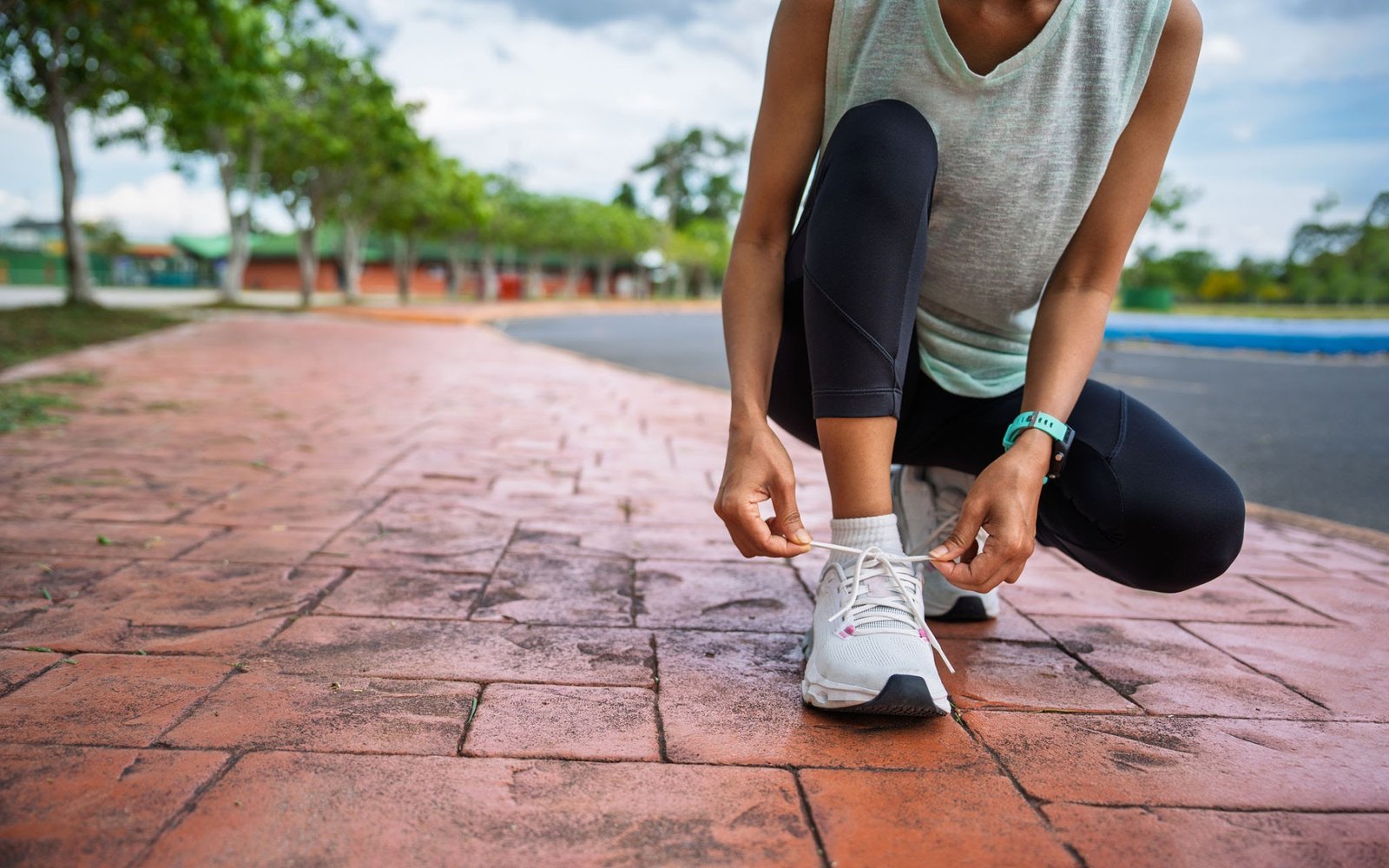 Identity-Based Habits Person tying white sneakers on a brick path outdoors, wearing a gray sleeveless top and black leggings.